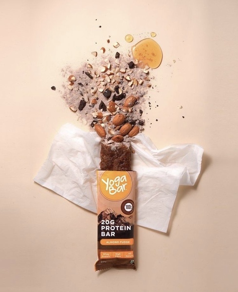 A top-down, minimalist product photography shot showing a Protein Bar surrounded by floating, deconstructed raw ingredients like almond fragments and syrup.